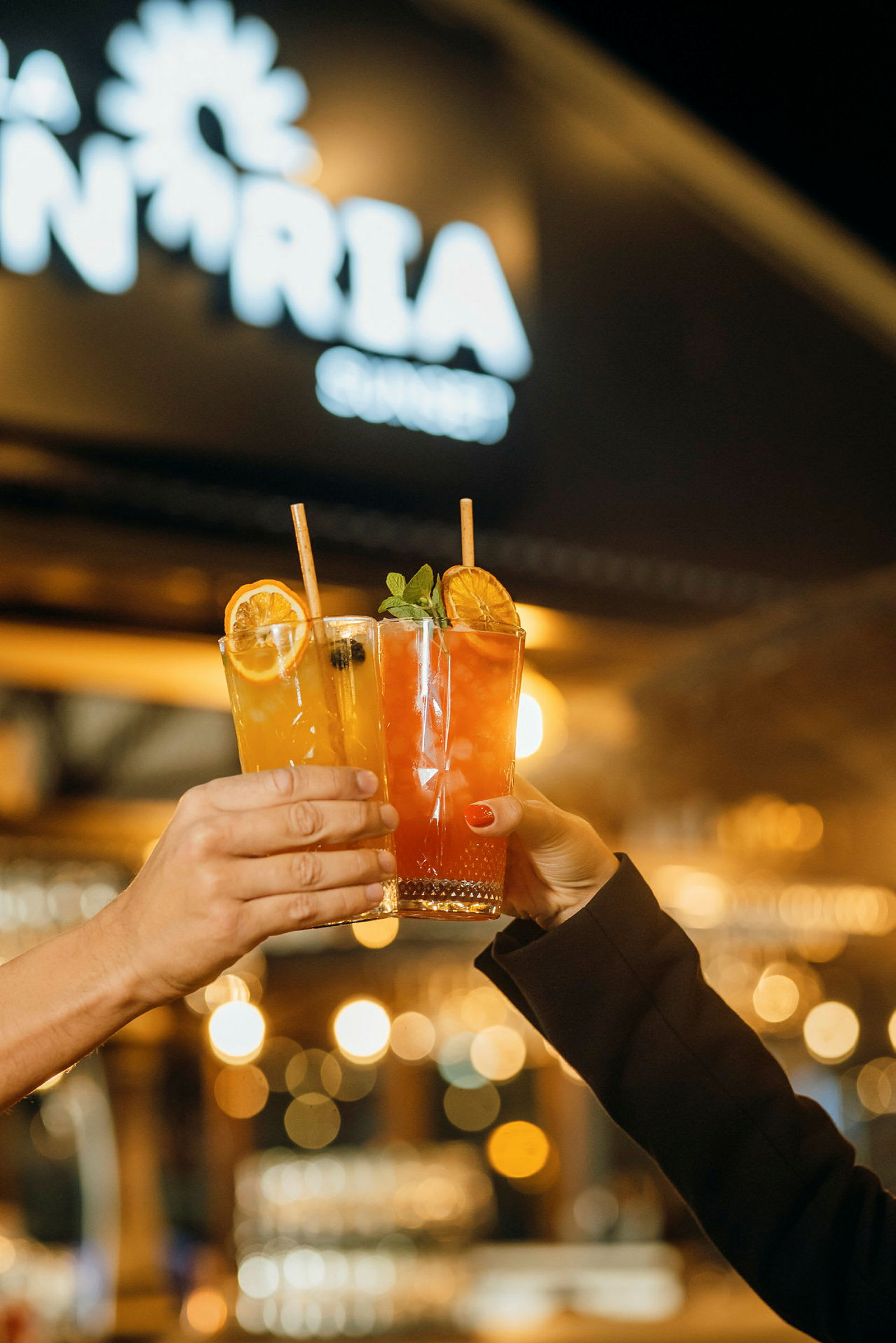 Två händer håller upp varsitt glas med färgglada drinkar framför en ljusskylt. (AI-genererad text)