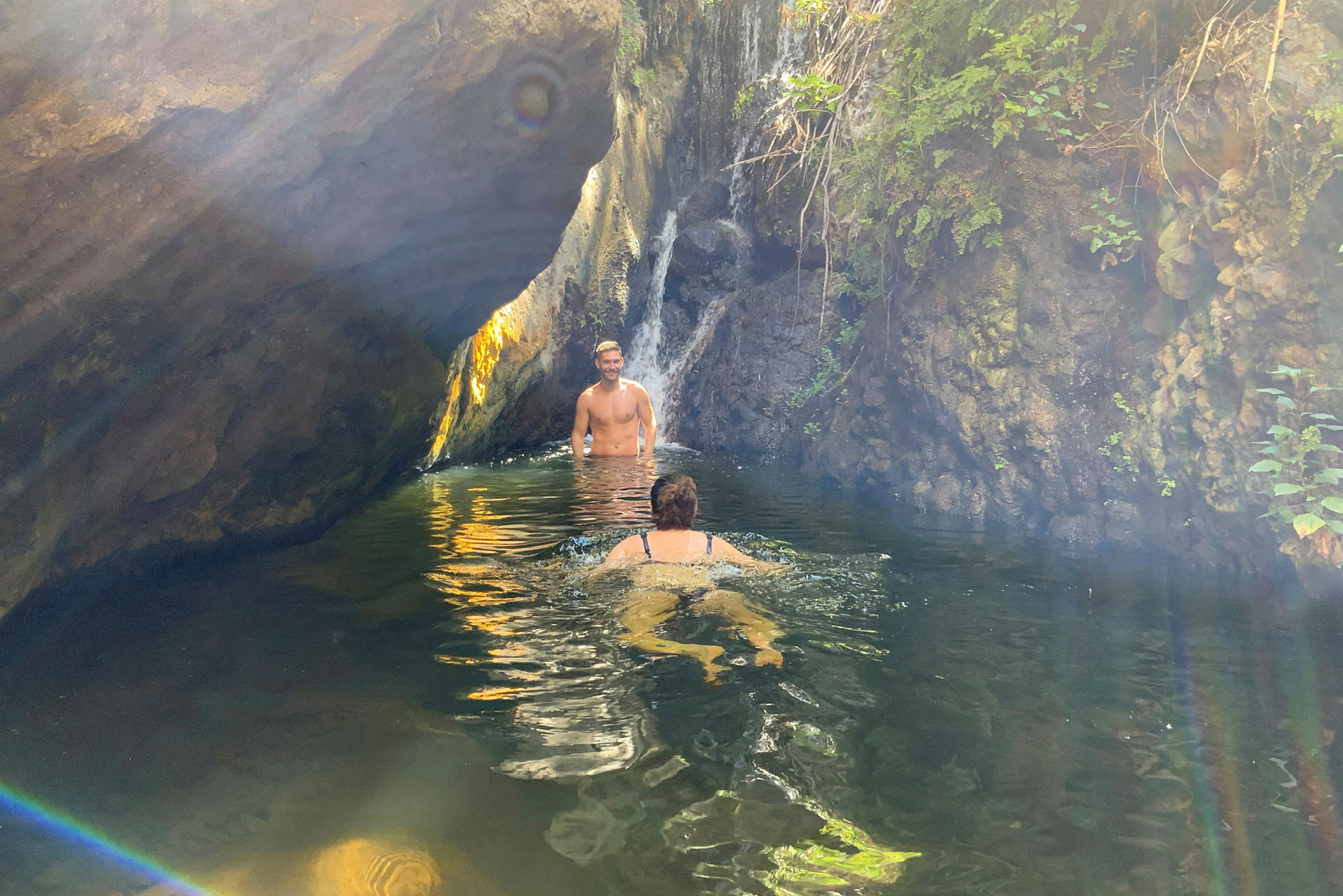 En person simmar i en naturlig pool intill en liten vattenfall bland klippor och grönska. (AI-genererad text)
