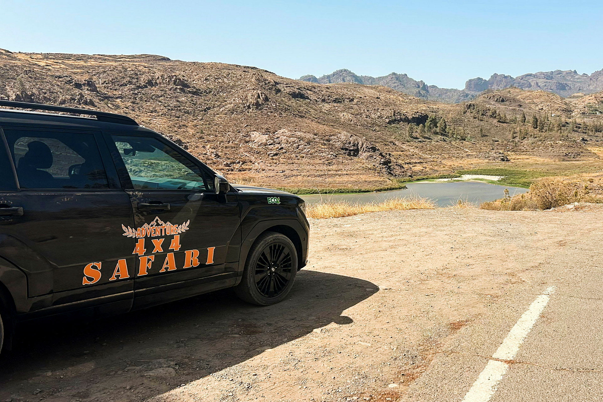 En fyrhjulsdriven bil med texten 'Adventure 4x4 Safari' parkerad i en ökenliknande miljö med en sjö i bakgrunden. (AI-genererad text)
