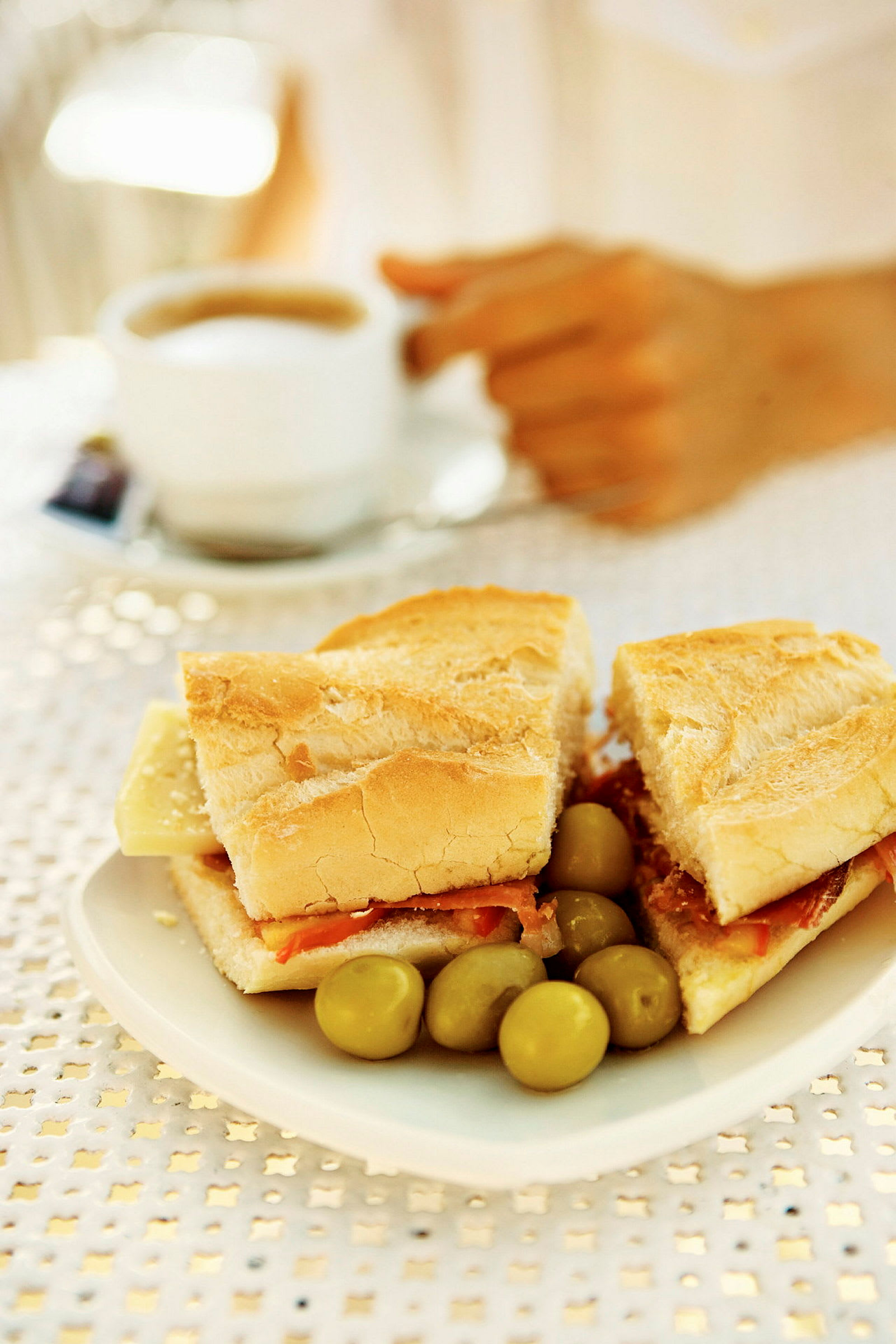 En tallrik med en baguette fylld med ost och skinka samt några oliver. I bakgrunden syns en hand som håller en kopp kaffe. (AI-genererad text)