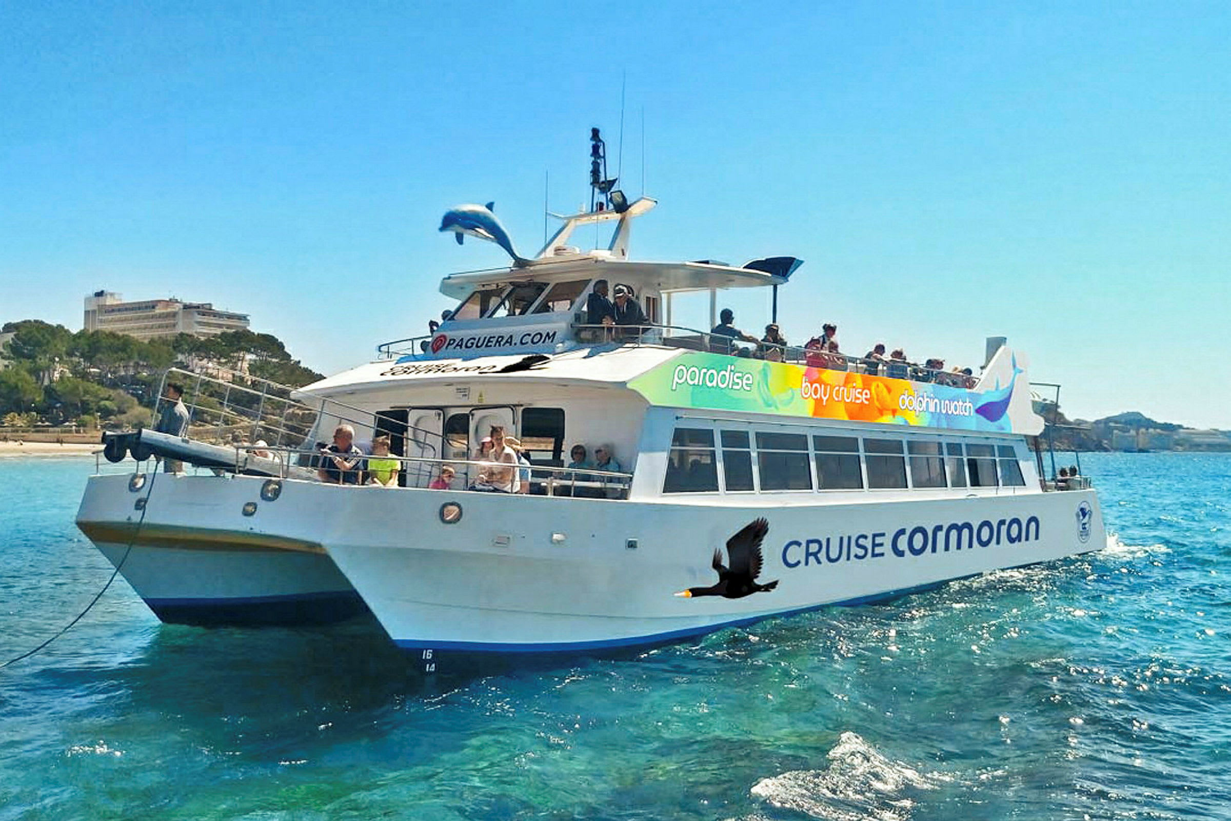 Ett fartyg med namnet 'Cruise Cormoran' på vattnet, omgiven av passagerare och med blå himmel och landskap i bakgrunden. (AI-genererad text)