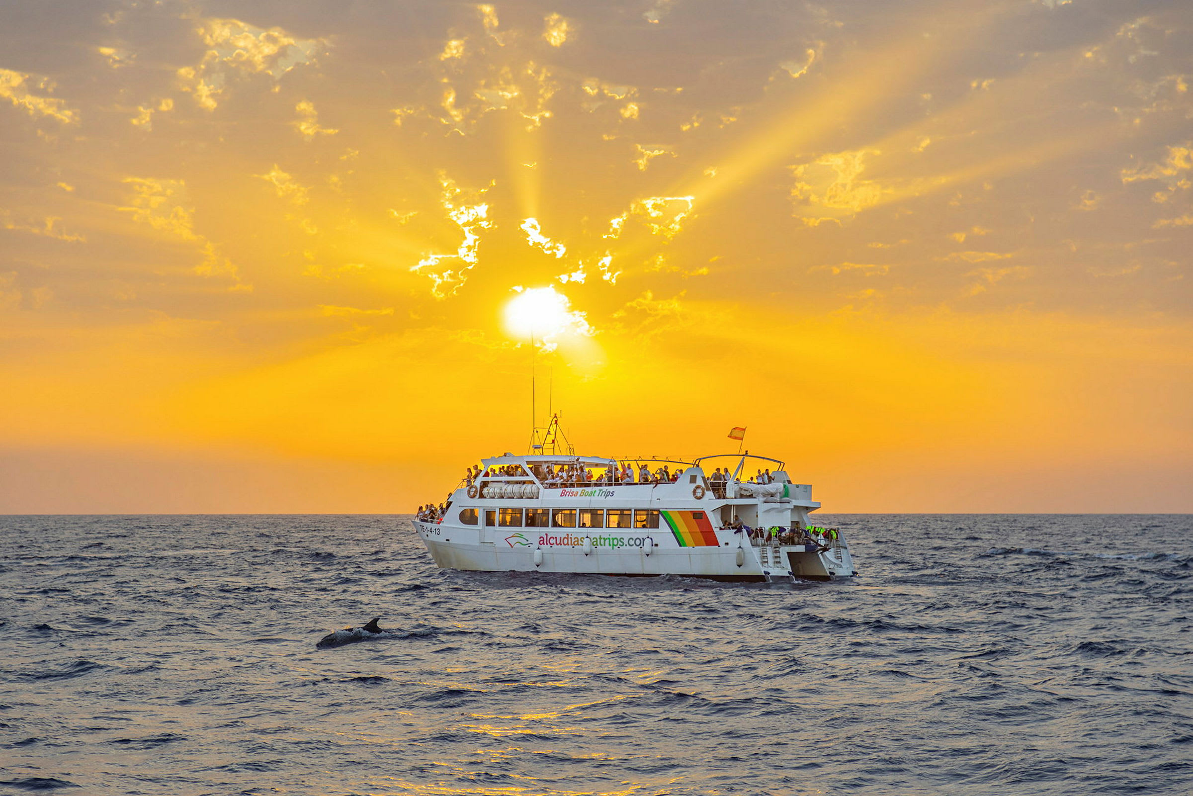 En vit båt med människor på däck flyter på havet under en orange solnedgång. (AI-genererad text)