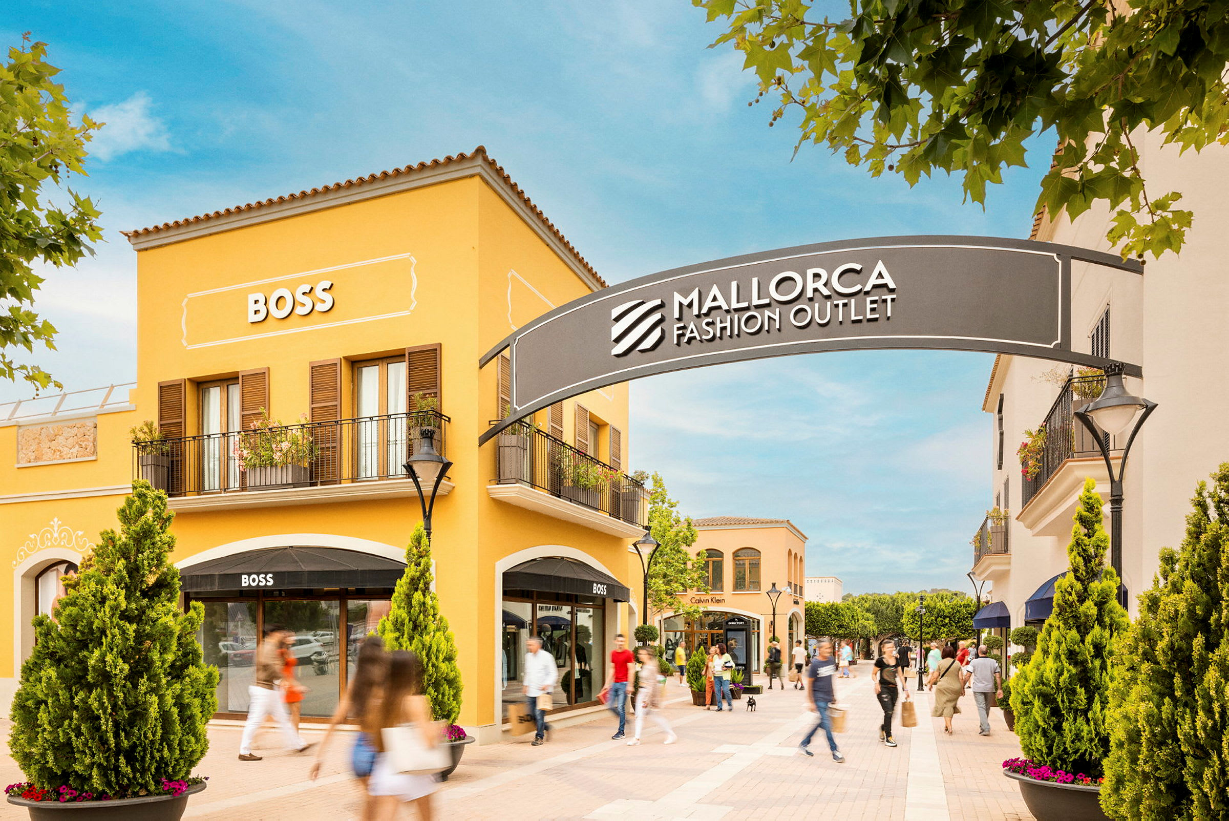 En shoppinggata med gula byggnader som visar skyltar för butiker, bland annat BOSS. Ett skylt med texten "Mallorca Fashion Outlet" syns ovanför ingången till området. (AI-genererad text)