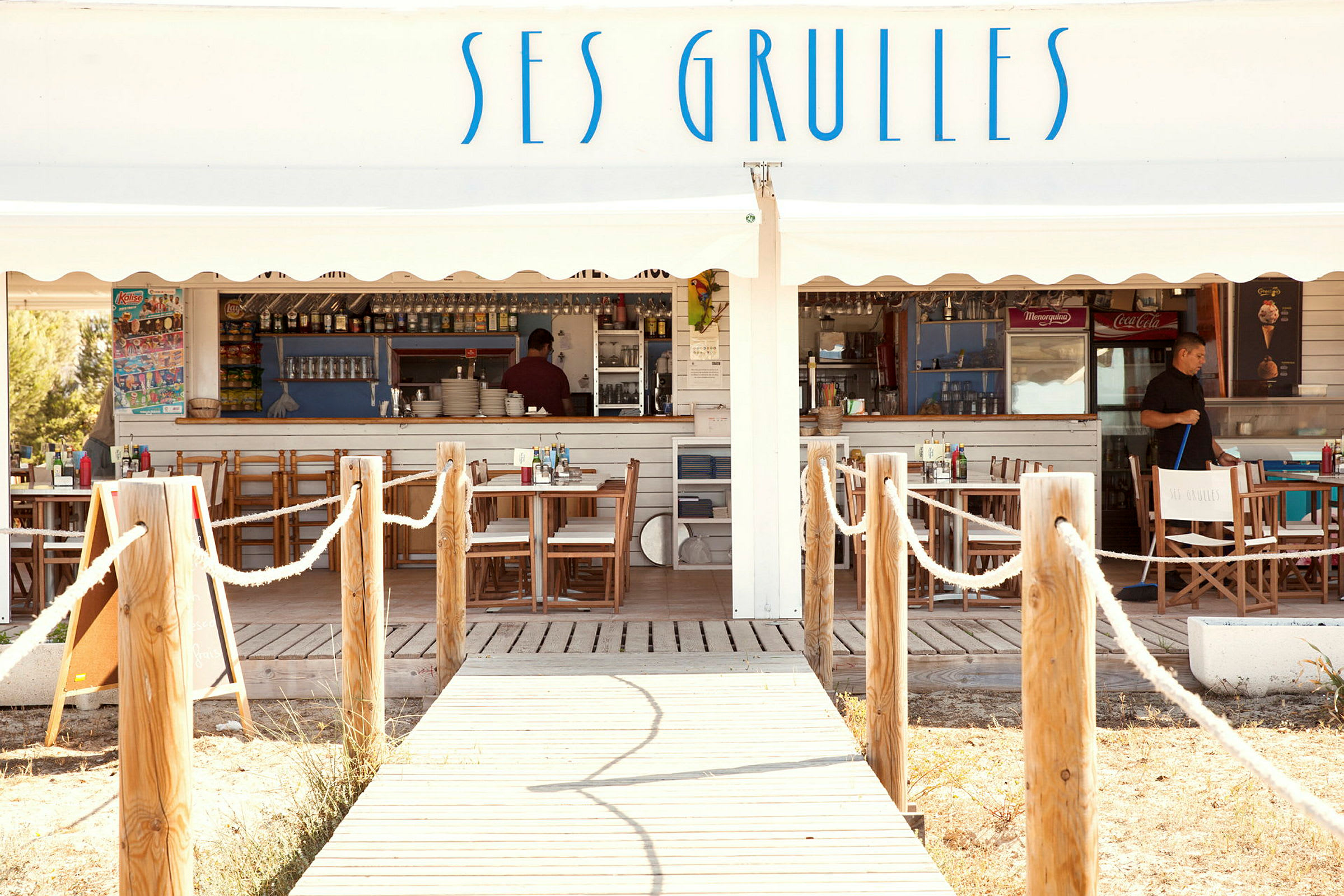 En trägång leder till en utomhusrestaurang med vita träväggar och skylttexten "Ses Grulles". (AI-genererad text)