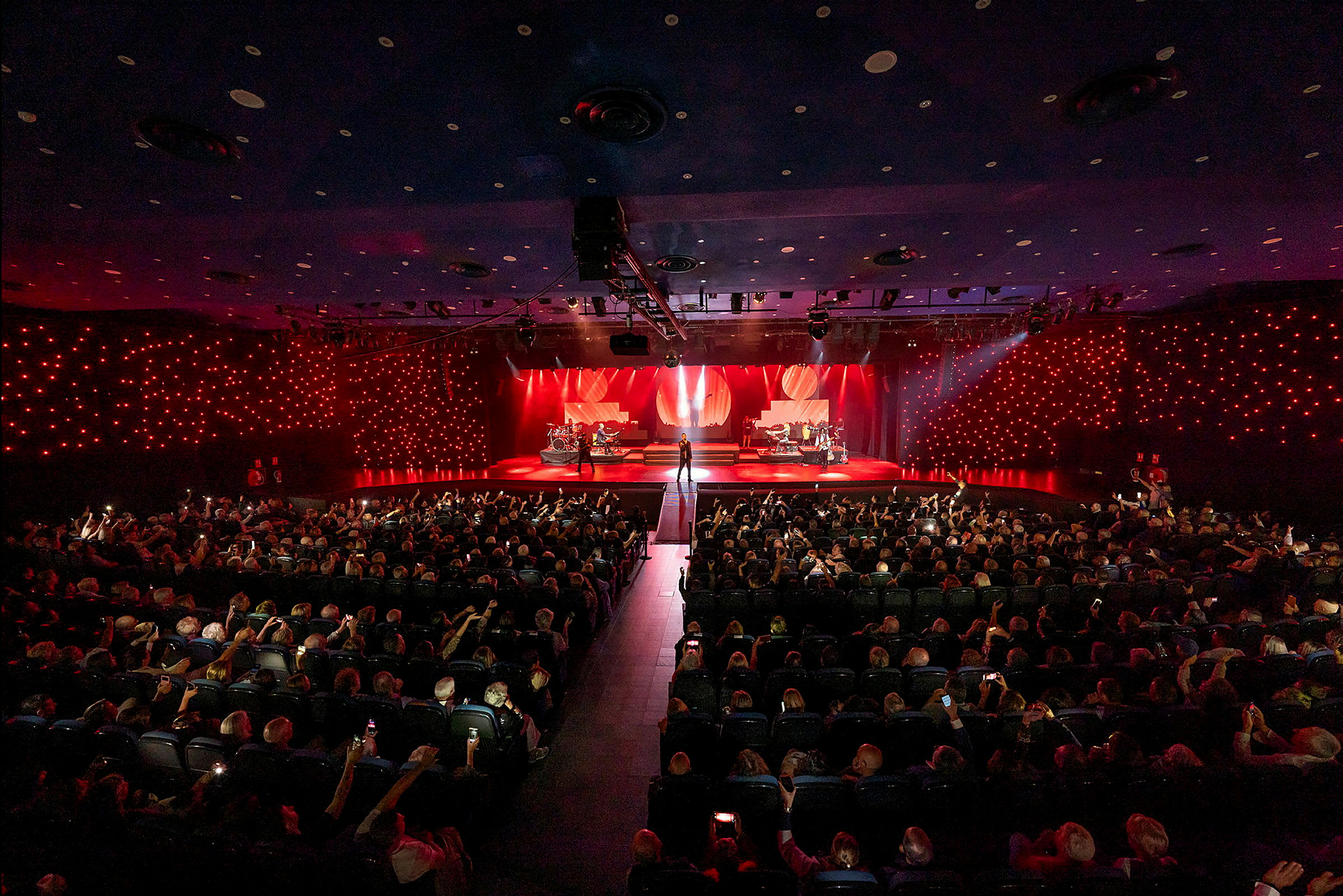 En konsertsal med en upplyst scen där olika musiker framträder inför en publik. (AI-genererad text)