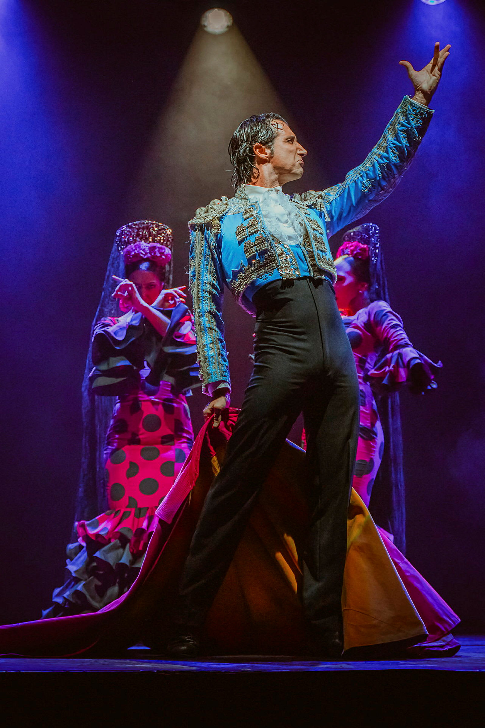 En flamencodansare i färgrik dräkt poserar på en scen med två bakgrundsdansare i traditionella kläder. (AI-genererad text)