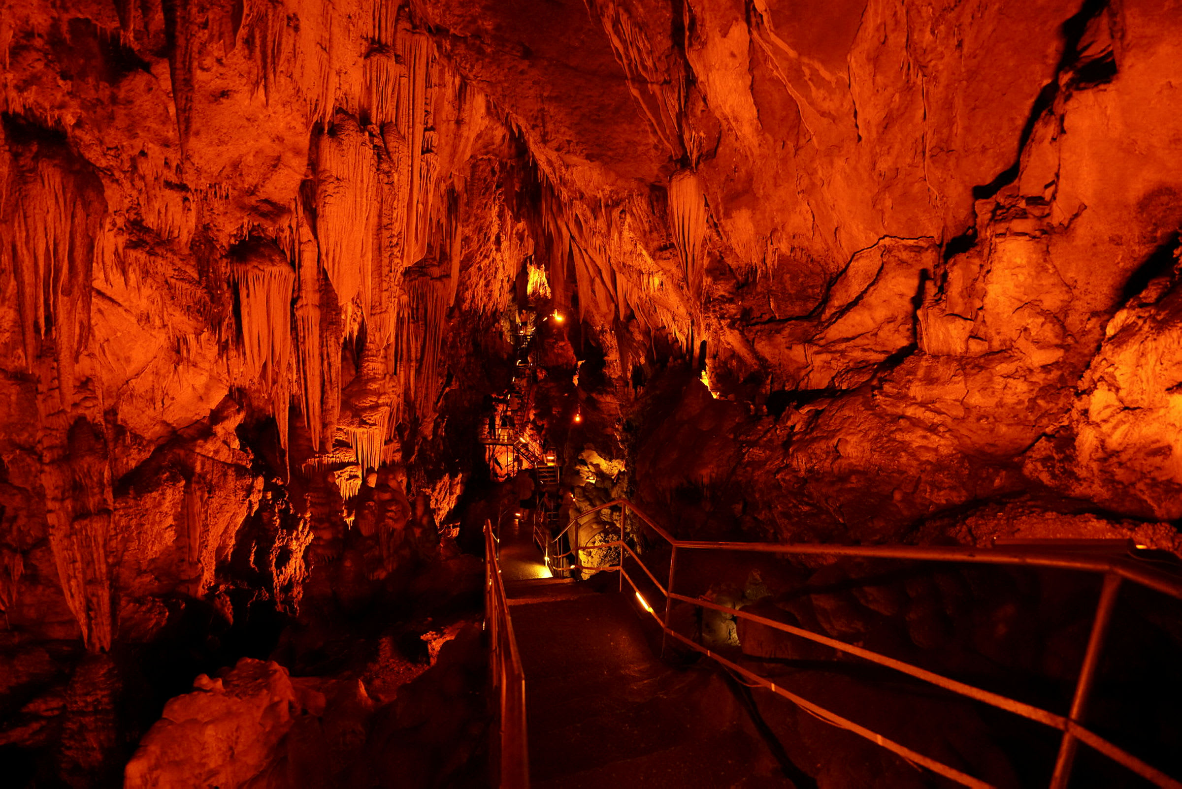 En grotta med stalaktiter och stalagmiter, upplysta med orange belysning. En gångväg med räcken leder genom grottan. (AI-genererad text)
