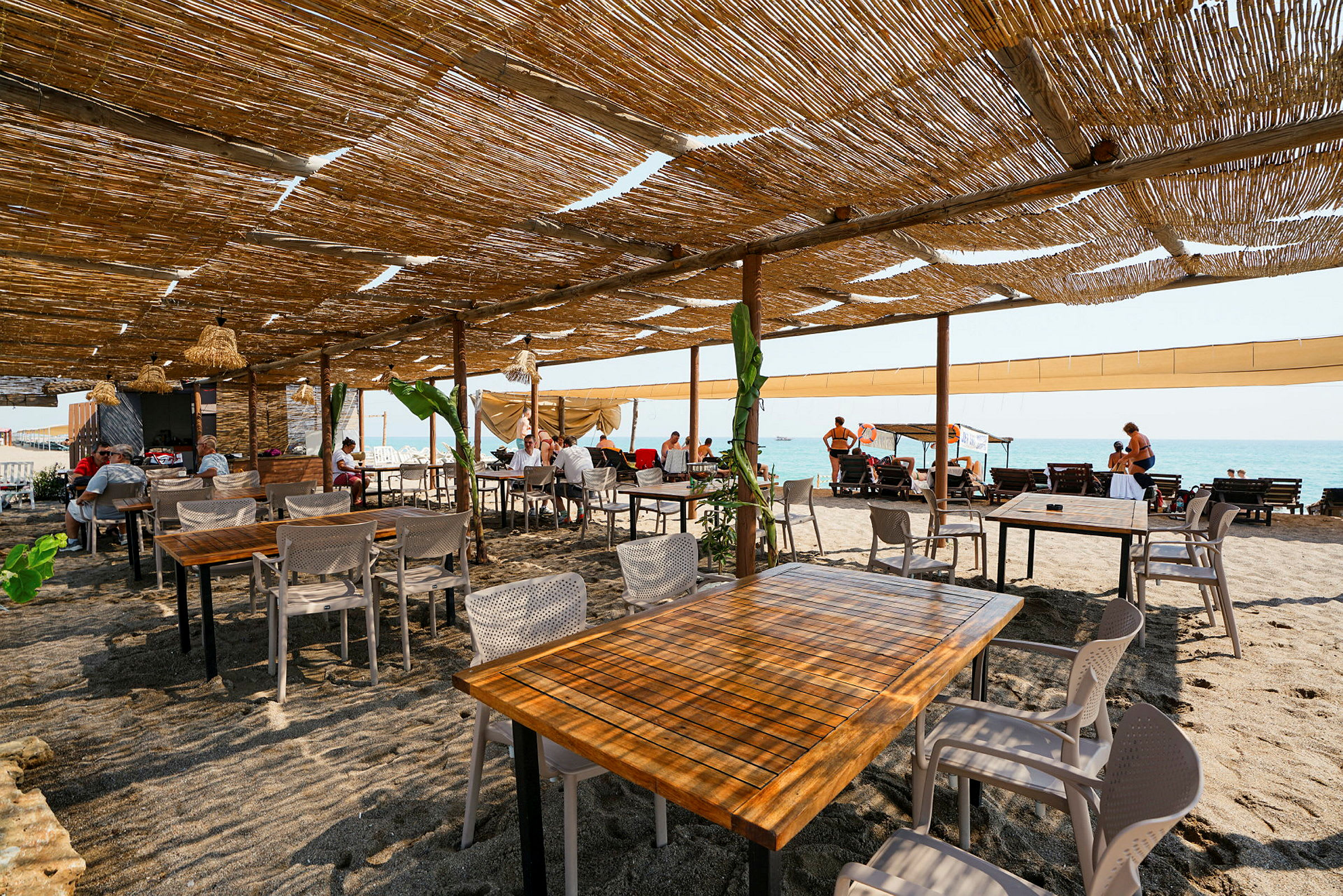 Utomhusrestaurang med bord och stolar på sandig strand under tak av bambu. (AI-genererad text)