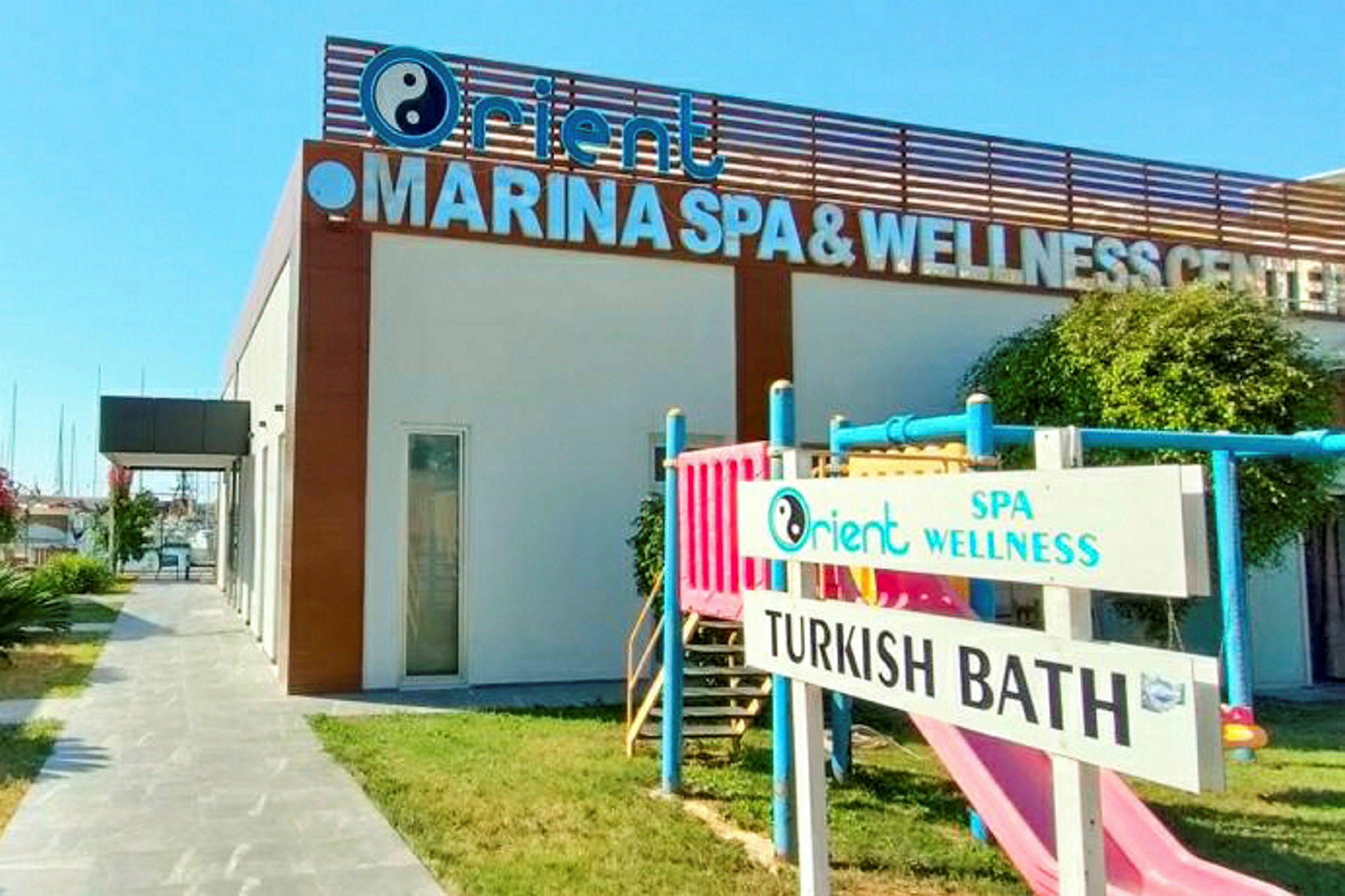 Byggnad med en skylt där det står 'MARINA SPA & WELLNESS CENTER'. På gräsmattan finns också en barnlekplats och en skylt med texten 'TURKISH BATH'. (AI-genererad text)