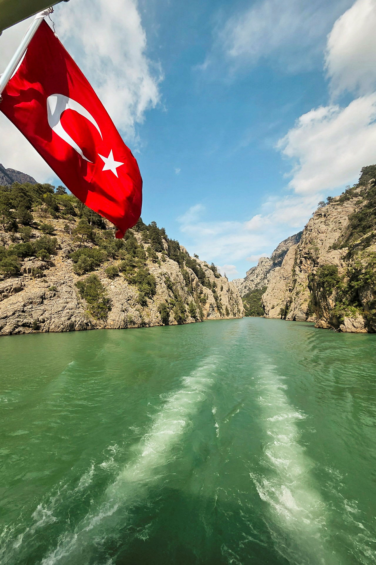 En vy från en båt som färdas mellan klippor i en grön flod, med en turkisk flagga i förgrunden. (AI-genererad text)