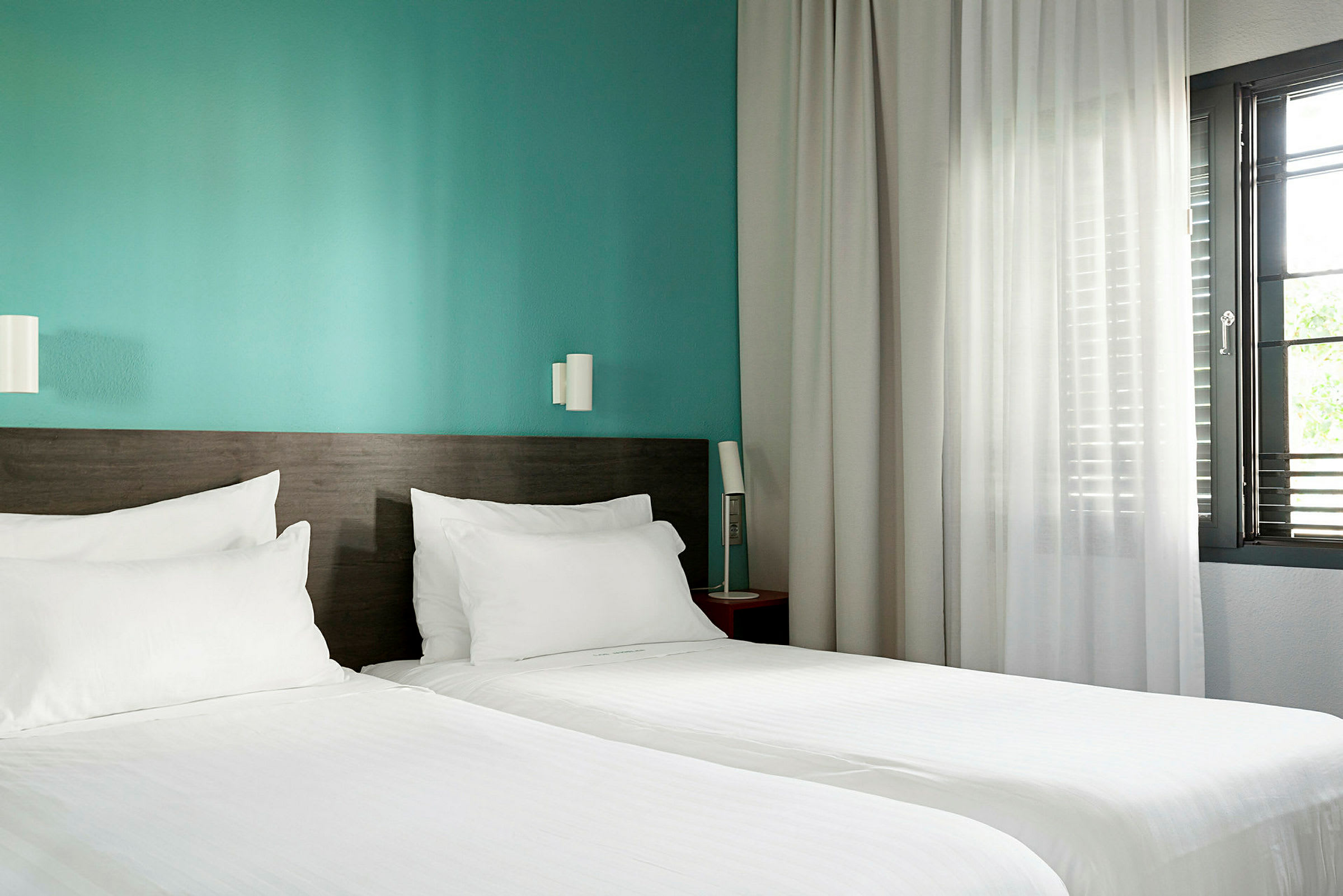 Ett hotellrum med två enkelsängar, en turkos vägg och ett fönster med vita gardiner. (AI-genererad text)
